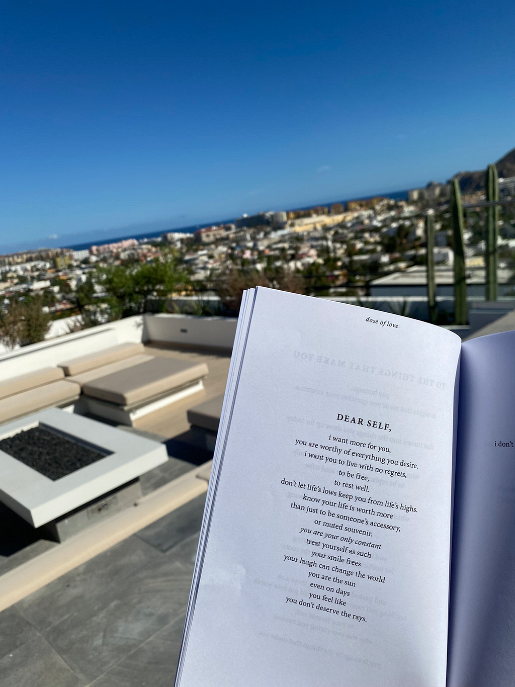 A book of poetry is opened up on a sunny rooftop patio. In the backround, a sunny, suburban cityscape with blue sky and an ocean view.