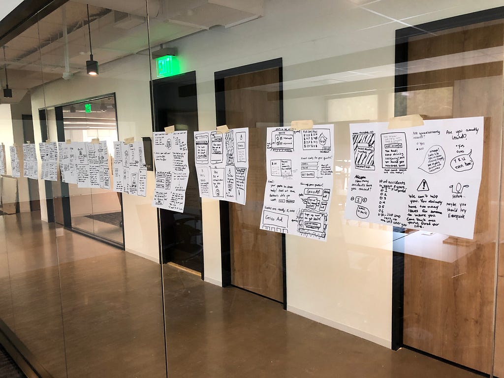 A row of hand-drawn sketches taped to a glass wall in an office hallway. Each sheet shows rough notes, doodles, and storyboard-style ideas created during a Black Mirror design exercise.