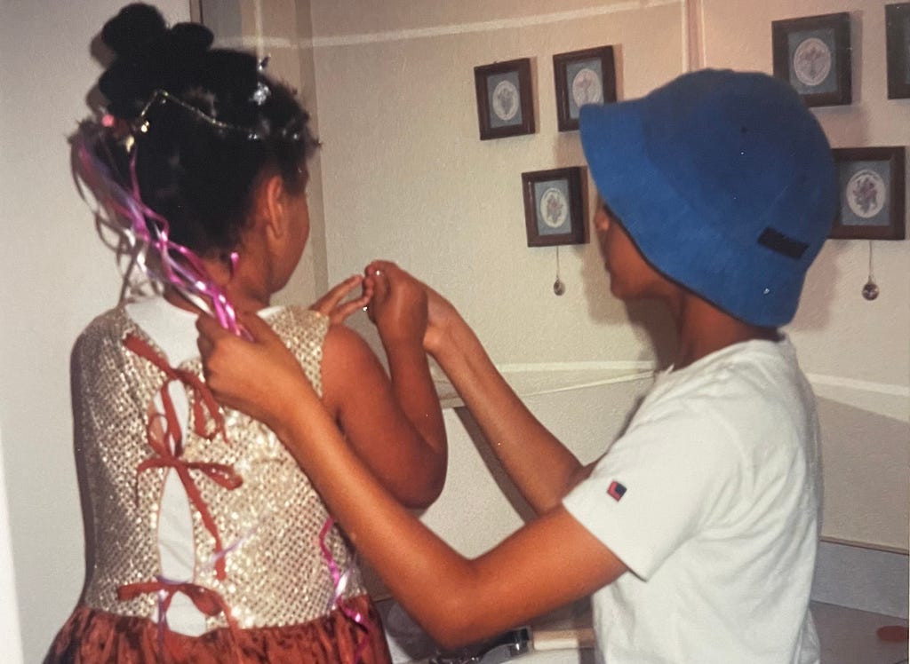 My older sister helping me dress up when I was around 7 and she was around 12, the picture is taken from behind us so you can see my hair covered in ribbons and we’re both holding onto something sparkly in our hands.