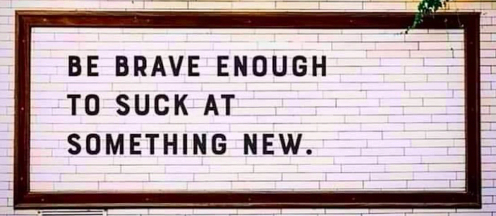“BE BRAVE ENOUGH TO SUCK AT SOMETHING NEW.” written in black all-caps text against a white brick wall.