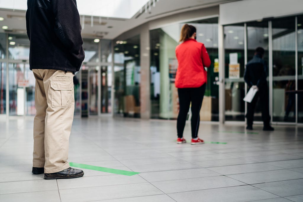 People queue, standing on lines taped to the floor to indicate a safe distance between them