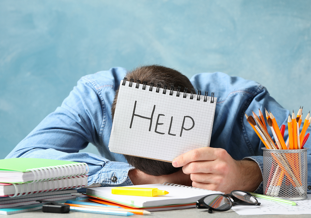 Man wearing a blue shirt with his head down on his desk. He’s holding up a notebook with the word “help”.