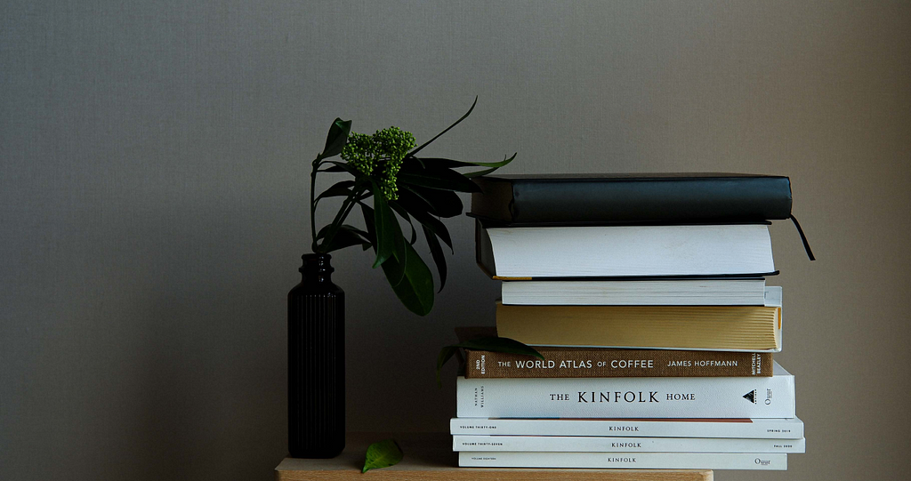 a pile of white, brown and black books on a small table or shelf (the actual object unseen) and a black vase with some dark green leaves inside standing next to it