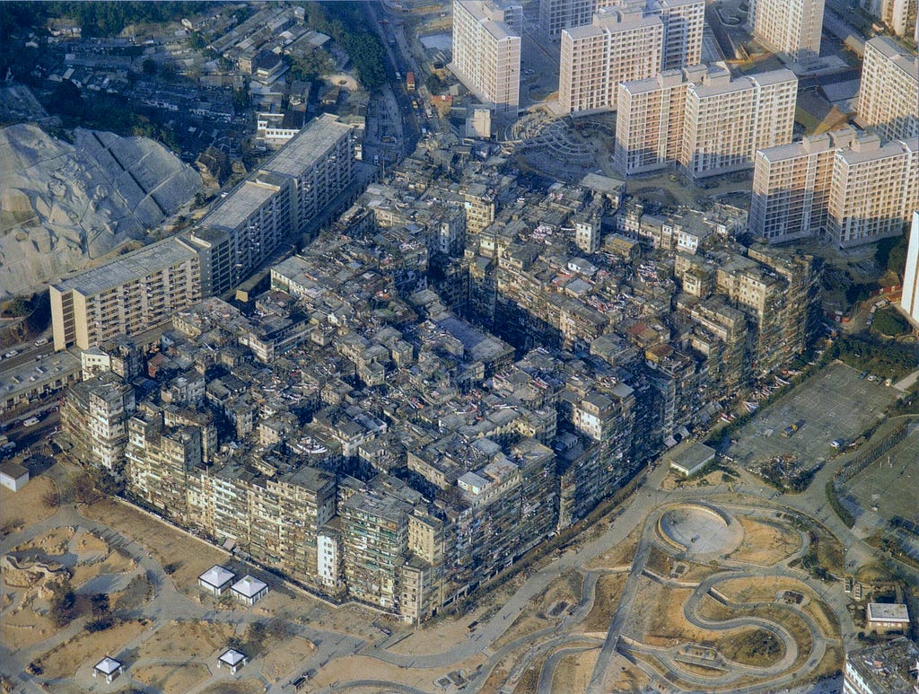Aerial photograph of Kowloon Walled City, Hong Kong, 1989, showing an extraordinarily dense cluster of tightly packed, low-rise buildings forming a single continuous urban mass, surrounded by modern high-rise apartment blocks, open land, and a curved road.