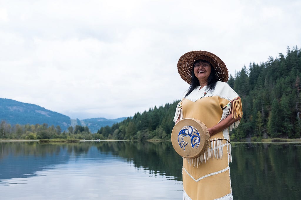 Salish coastal Native American woman stands proudly in traditional clothing before a Puget Sound background of inner-coastal water, trees, and rolling foothills.