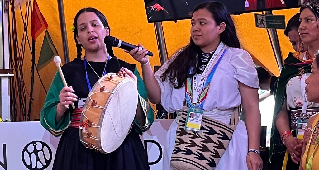 A person holding and playing a tambor drum and singing. Another person next to them is holding a microphone up to capture their performance