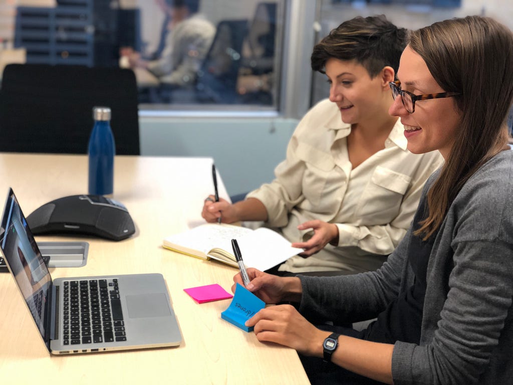 Two women are conducting a remote interview with a drone pilot. They sit smiling and looking towards a laptop, while taking notes on post-its.