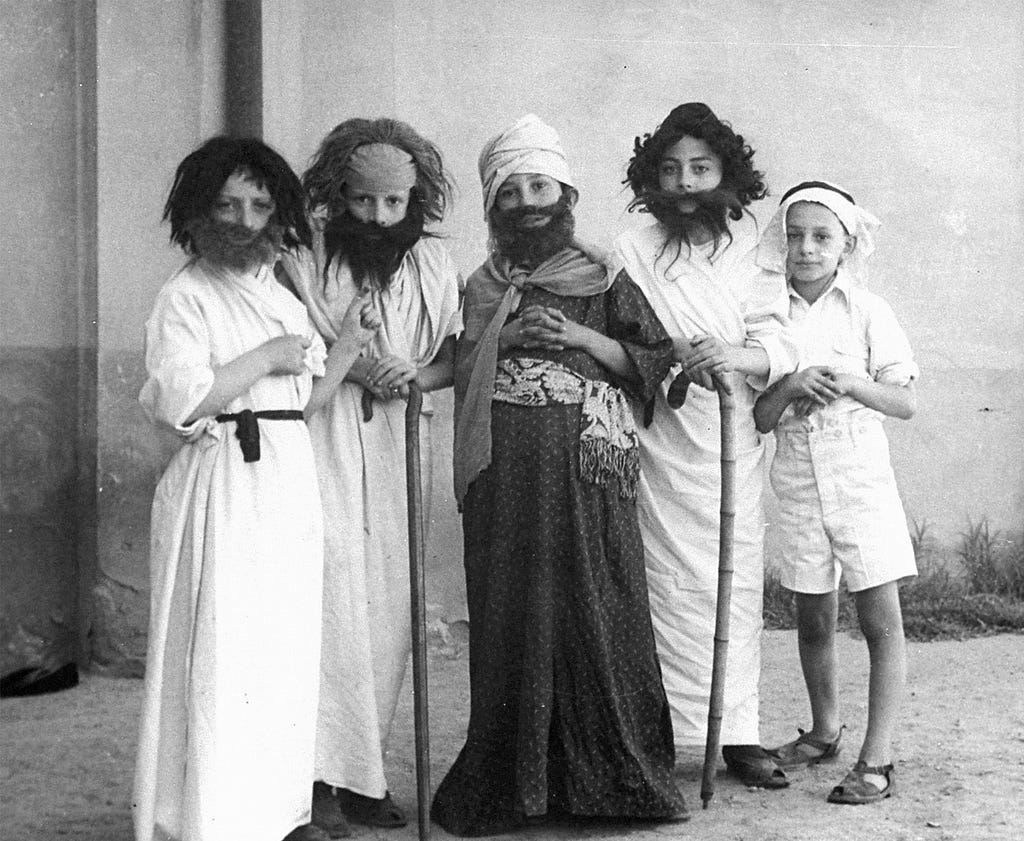 Five boys dressed in biblical costumes standing in a row outside in front of a blank wall. The ground is covered in dirt. Four are wearing wigs and fake beards. Two hold canes. The boy in the center stands out, dressed in a patterned, dark full-length garment, while the others are dressed in solid, white full-length garments. One boy on the far right is wearing white shorts and sandals.