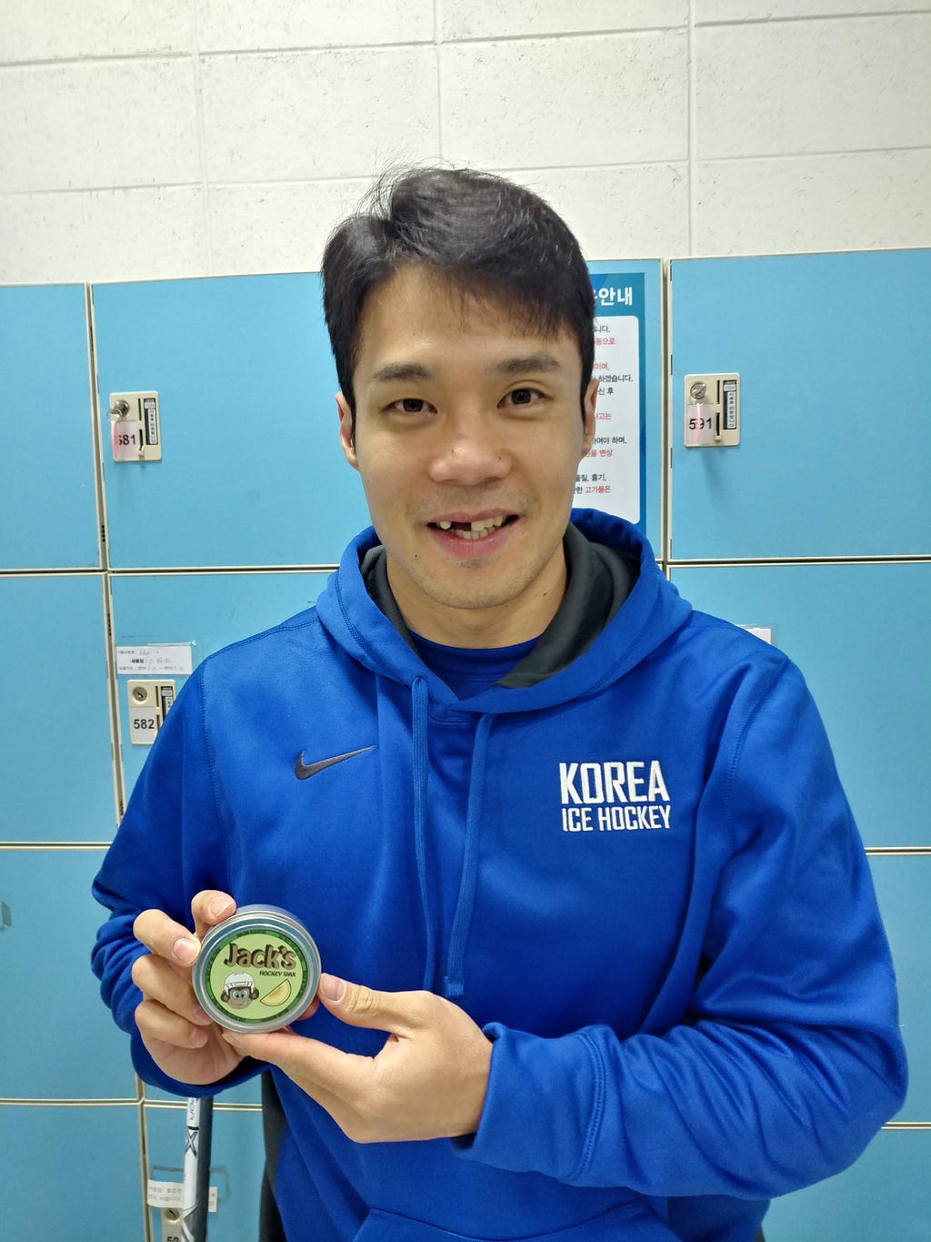 Korean ice hockey player Cho Minho holds a tin of Jack’s Hockey Wax while smiling in front of a wall of lockers.