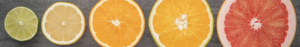Five cross sections of citrus fruits arranged in ascending order of diameter on a textured grey background. From left to right, they appear to be a lime, a lemon, and orange, a larger orange citrus fruit, and a red grapefruit.