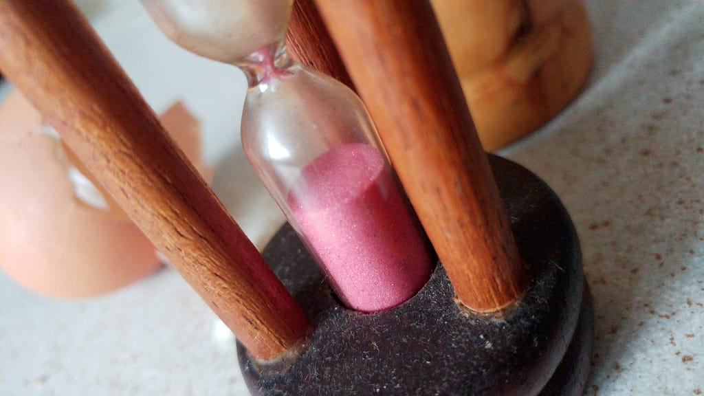 Photo of a wooden egg-timer with pink sand in a tiny hourglass. In the background there’s half an eggshell and a huge wooden pepper grinder.