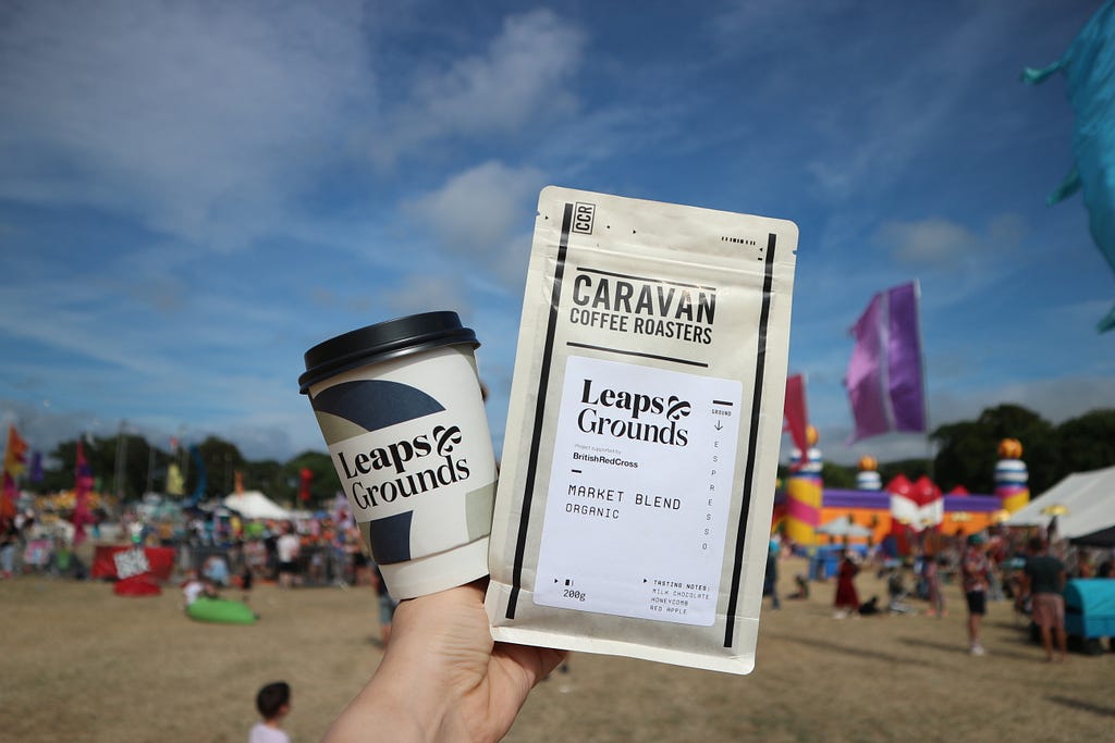 A Leaps & Grounds coffee cup alongside Leaps & Grounds/Caravan co-branded coffee bags, with a festival in the background