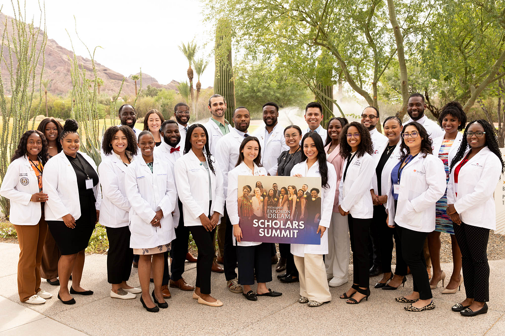 A large, diverse group of young professionals in white lab coats pose outdoors in a sunny desert landscape with cacti and trees. They stand together, smiling, while two people in front hold a sign reading &ldquo;Explosion Dream Scholars Summit.&rdquo; The group includes men and women of various ethnicities, dressed in professional attire.