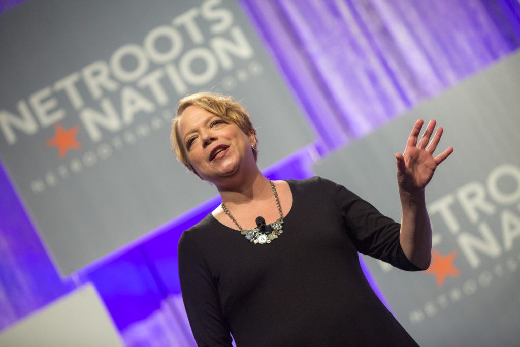 Photo of a white woman with short blonde hair, presenting on a stage with the words “Netroots Nation” behind her.