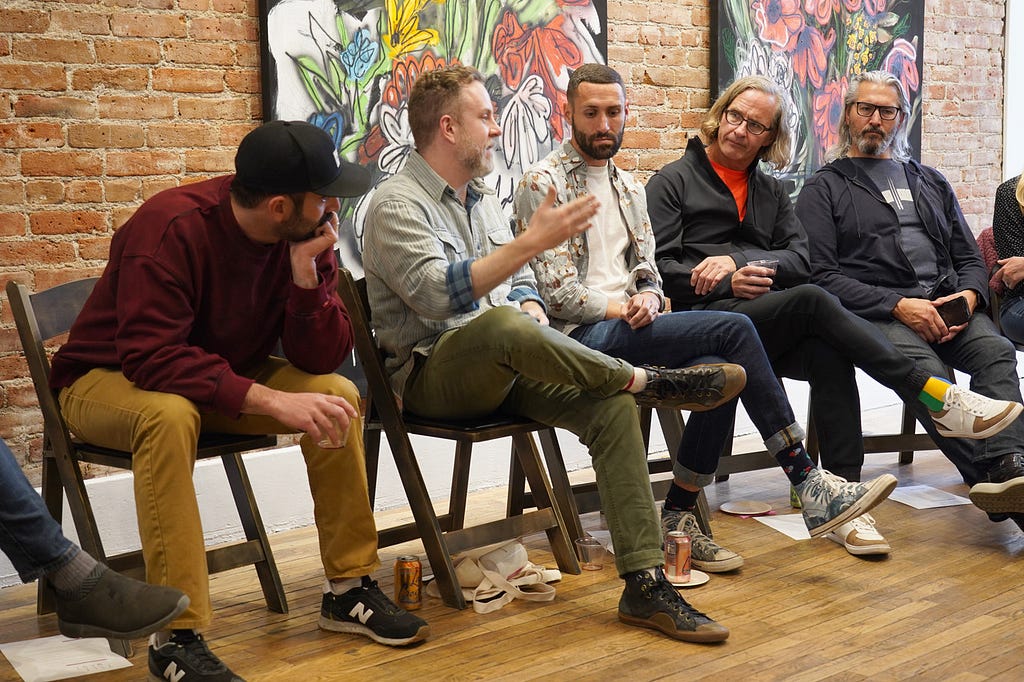 Five men sit in a line of black chairs flanked by large colorful paintings. One man speaks, his hand held forward in emphasis, while the others listen attentively.