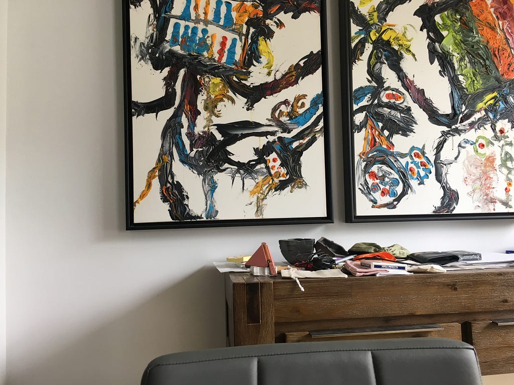 Two frames of an abstract painting hang above a cluttered hardwood console table. The top of a grey chair sits in front.