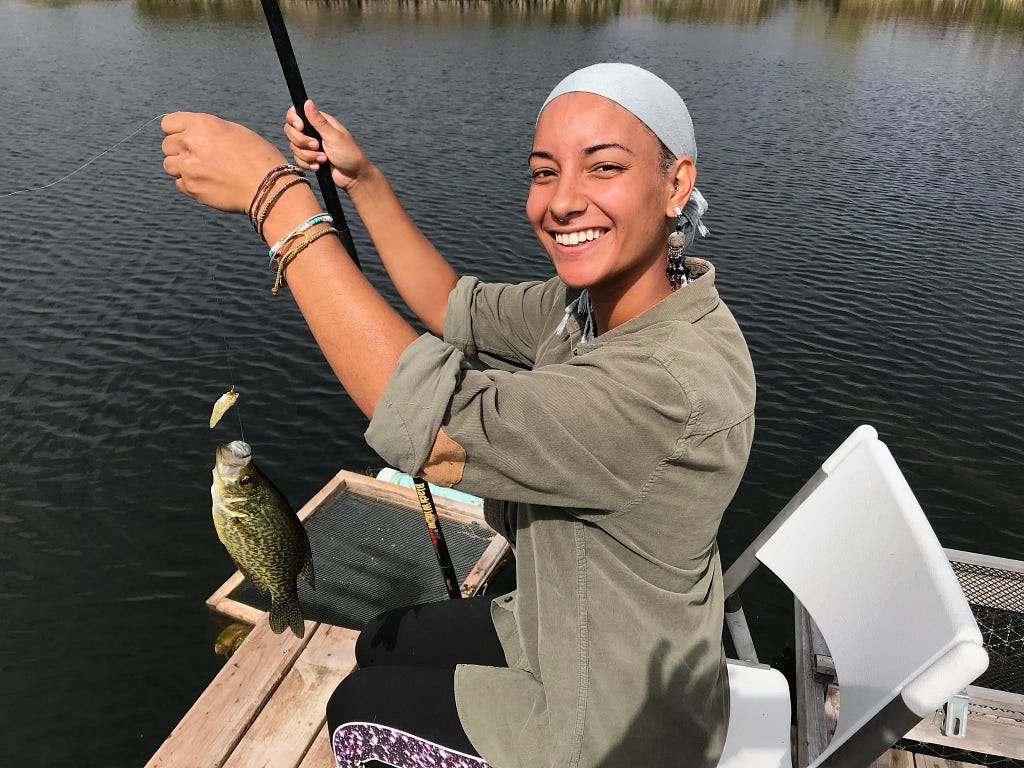 Young person of color smiling, sitting on a dock holding a fishing line with a fish she caught on the hook