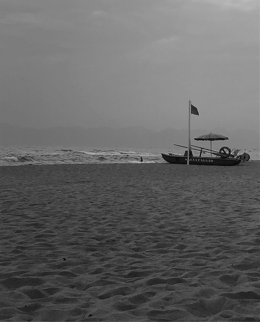 A black and white picture of a beach. No people can be seen, just a hissed flag and a safe boat reading ‘Salvataggio’.