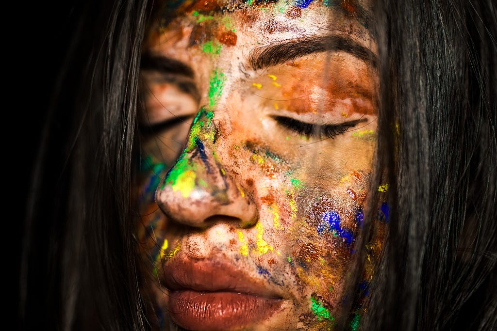 A caucasion woman’s face covered with streaks of paint in different colours.