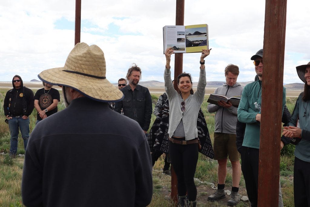 Mercedes Martinez holds the LAGI 2020 Fly Ranch book over her head, open to the page about “The Loop.” Around her are ten others with many more outside the picture frame. Tall poles mark the boundary of the project to help visualize what it will look like when built.