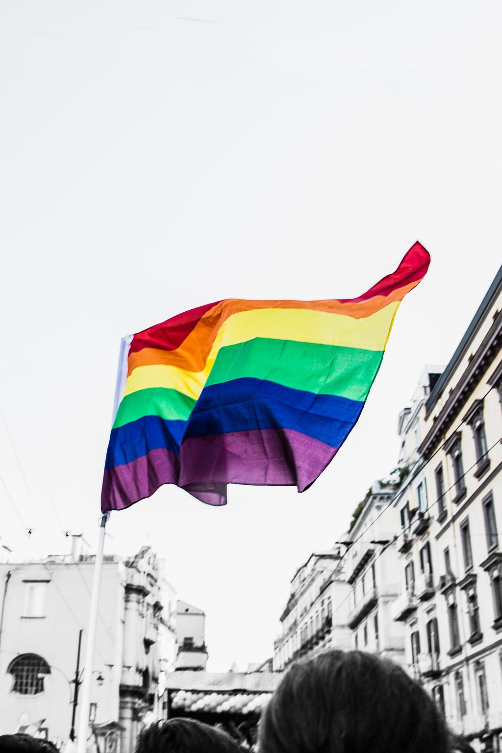 Imgem de bandeira LGBT+ em destaque no céu. É possível a cidade ao fundo, com prédios antigos.