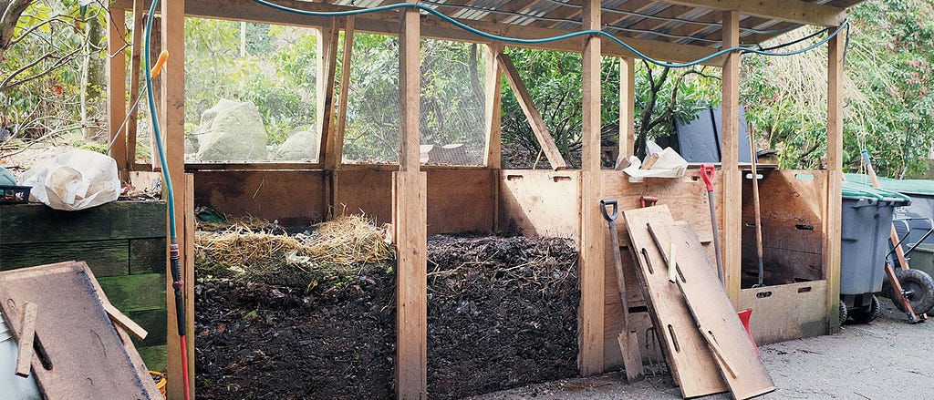 Four piles of dark soil compost, two of them in 1-metre-high piles, in a wooden shed-like structure. Behind the compost piles are a lush garden of vegetables and trees.