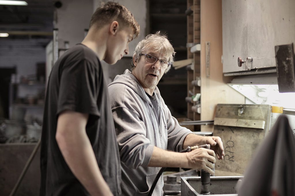 Free Side view of senior foreman in eyeglasses showing to trainee how handling detail in workshop Stock Photo