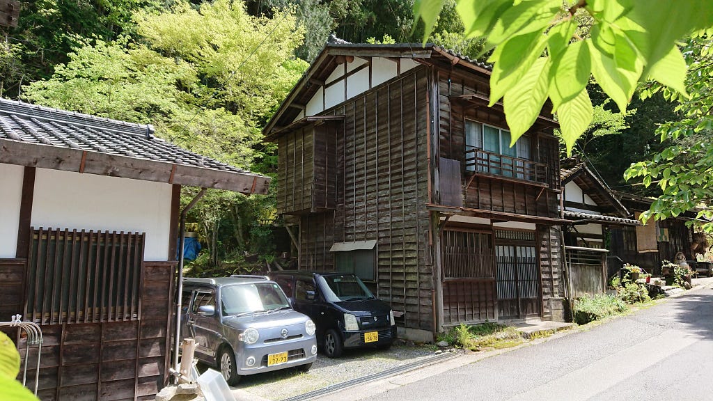 Two little cars sit in the drive of a beautiful dark-timber house on a leafy slope