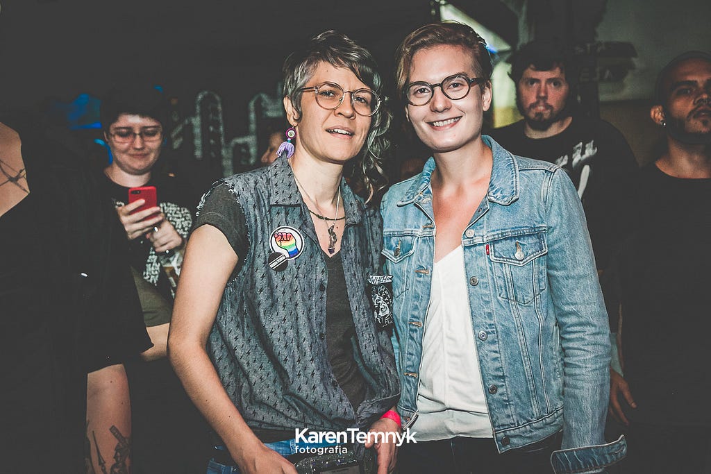 Puni e Gisele estão lado a lado com diferentes pessoas ao fundo, como em um show. Vestem jaquetas jeans com bottons arco-íris.