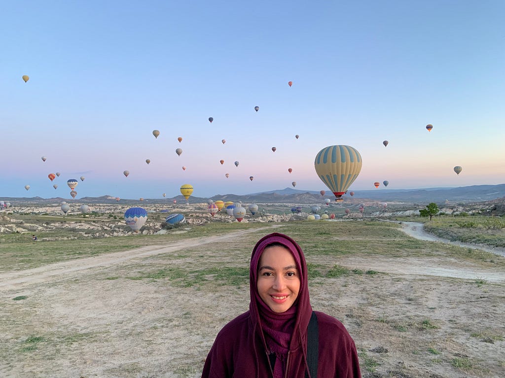 Anisa stands in the forefront wearing a maroon-colored hijab and a smile as dozens of hot air balloons fill the background against a sky at dusk.
