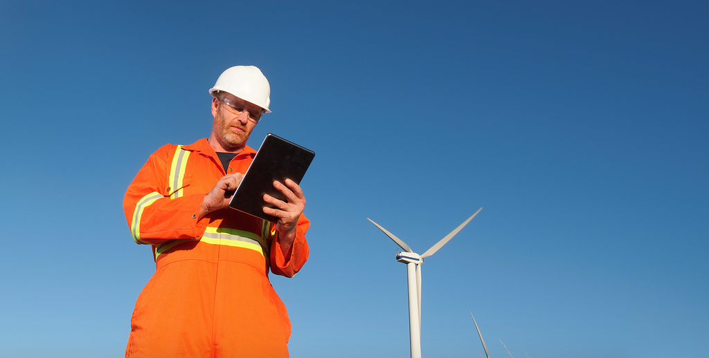 a person in an orange safety work outfit, hardhat, and goggles holding a tablet standing in front of a wind power windmill