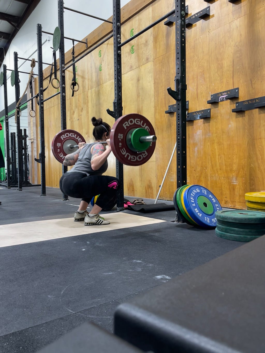 The writer, Kelsey, a white woman, is shown in the bottom of a back squat at a squate rack in a CrossFit gym. She’s wearing weightlifting shoes, leggings, a tank top, and an Aura 3M respirator.