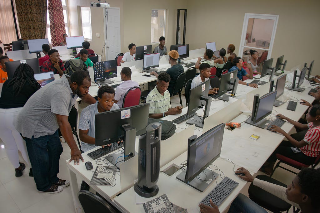 College students sit on front of several tables filled with computer monitors in a large classroom in Haiti.