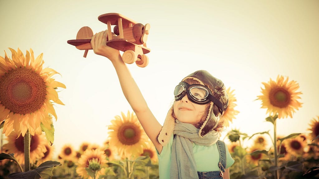 A child, wearing a pilot hat and glasses, plays with a toy airplane amidst a sunflower field.