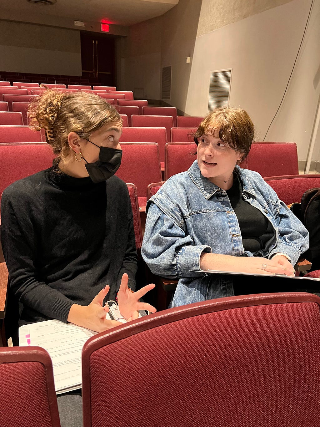 Two theater students sit in the audience of the theater talking to each other about rehearsals.