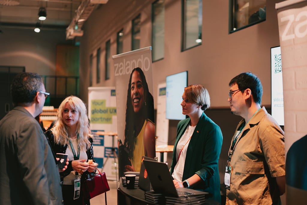 Four people standing around a booth and having a conversation, looking focused and listening attentively, with a Zalando-branded banner in the background.
