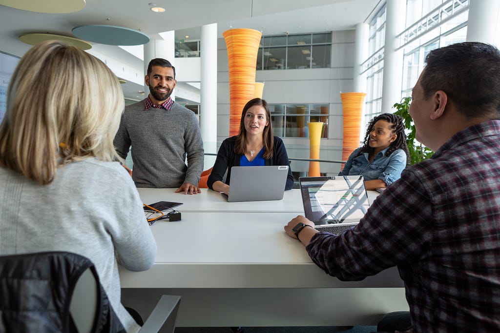 A group of five colleagues sit around a table in an office with open laptops, collaborating and talking