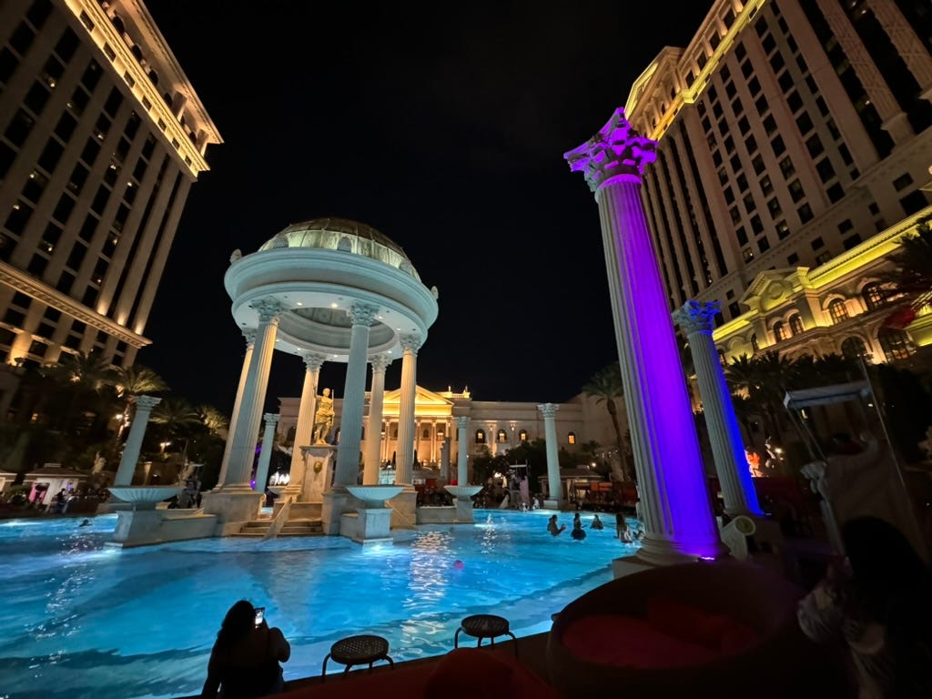A round circular pool with a Roman gazebo and purple lit pillars encircling the pool. Five attendees swim the center.