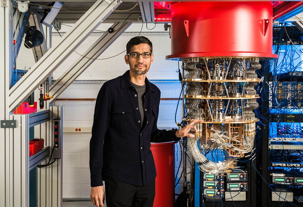 Sundar Pichai, CEO Google, stands next to a large, complex piece of quantum computing equipment. The person is wearing glasses and a dark jacket, and is interacting with the machine, which has numerous wires and components visible. The environment appears to be a high-tech lab, with various other equipment and cables in the background. The machine has a prominent red component at the top, and the overall setting suggests advanced technological research.