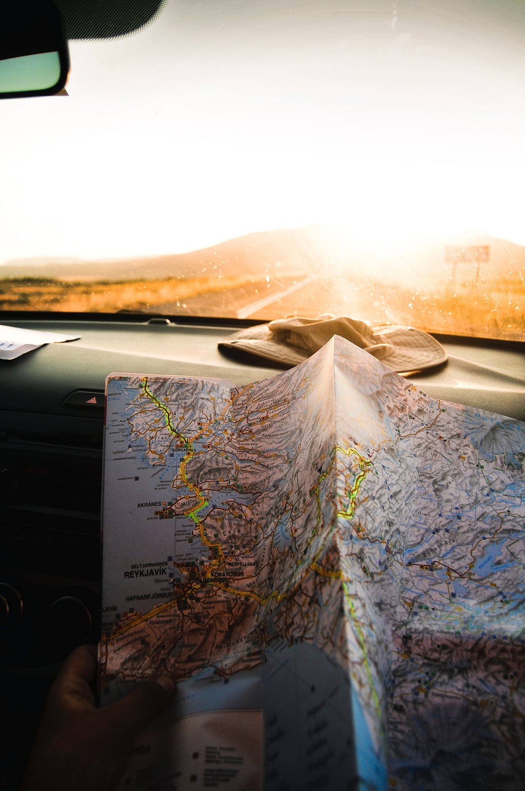 A map rests against a car dashboard as a sunset is visible through the windshield. Photo by Julentto Photography on Unsplash