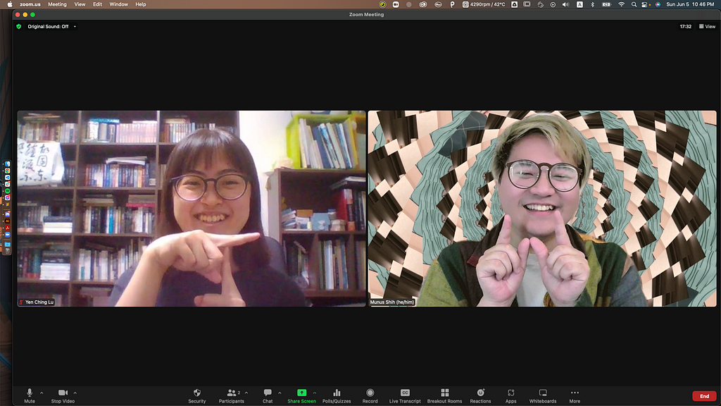 A screenshot of a Mac computer with a fullscreen Zoom meeting window. The window is divided into two camera screens with Yenching on the left and Munus on the right. Yenching has straight bob hair and wears round glasses and an indigo t-shirt. She is sitting in her study room and posing a letter T with her hands, smiling at the camera. Munus has bleached hair with a curvy bang and wears round glasses and a warm color block patched shirt. He is sitting with a kaleidoscope zoom background, and pos
