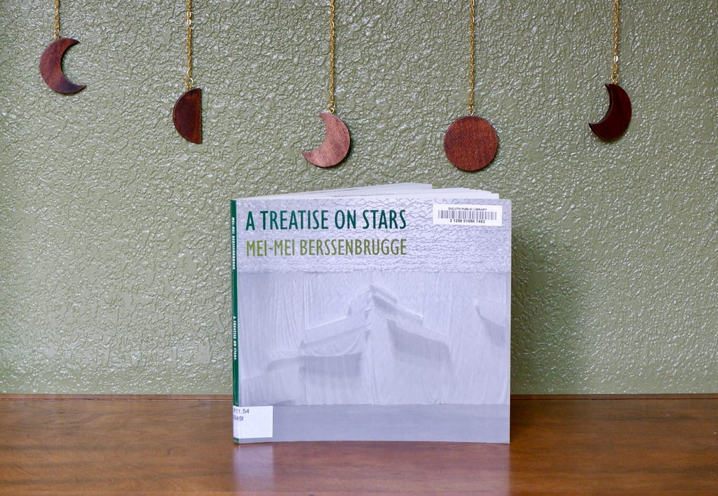 A book with a white cover and green lettering stands on a wooden shelf. Behind the book, a wooden moon phase garland is strung along a green wall.