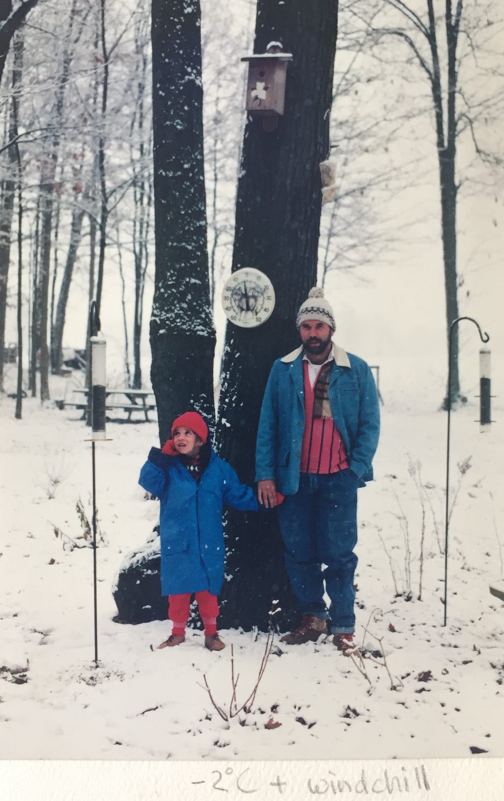 a small child in an over-sized winter coat holds hands with her father, both wearing knitted hats. There is snow on the ground and trees behind them. A hand-written note at the bottom of the photo reads “-2 Celcius + windchill”