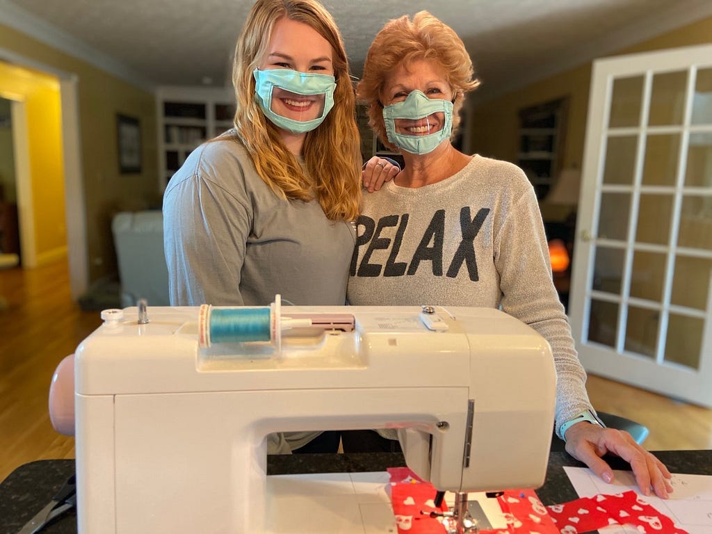 Two white women with masks with clear plastic standing behind a sewing machine.