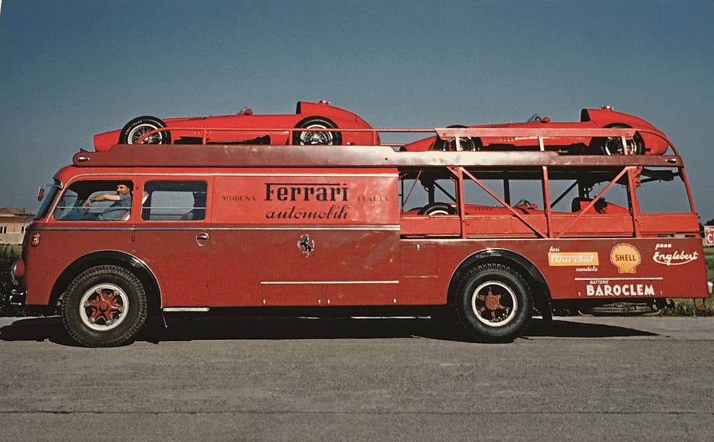 A red bus bearing the words “Ferrari automobiles” carries two racecars on the top (also red). A driver, his shirt the same blue as the sky, looks out the window disaffectedly.