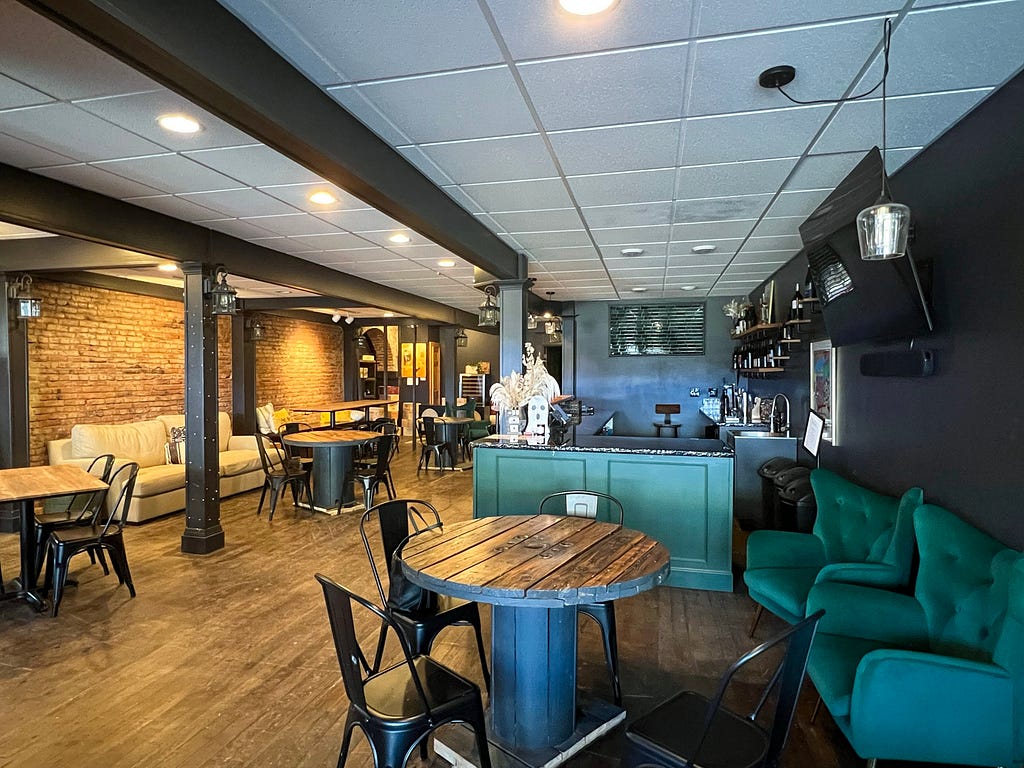 A cream colored couch sits against the brick wall lining the left side of the interior of a wine bar, The Cellar. The other walls and beams are black with a dark green bar and plush chairs. Wood tables with black chairs are visible throughout. A large television and shelves with wine bottles are visible on the right side.