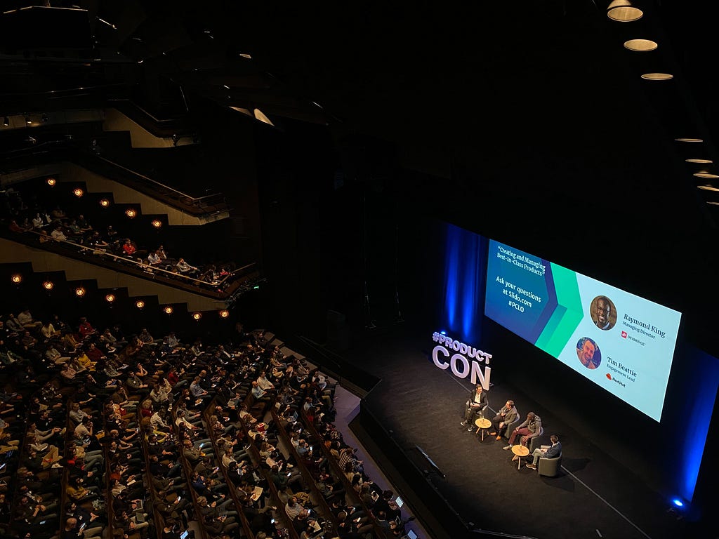 A picture of ProductCon Conference showing a stage with a few speakers on it and a powerpoint presentation in the background, before the stage many people are sitting on chairs.