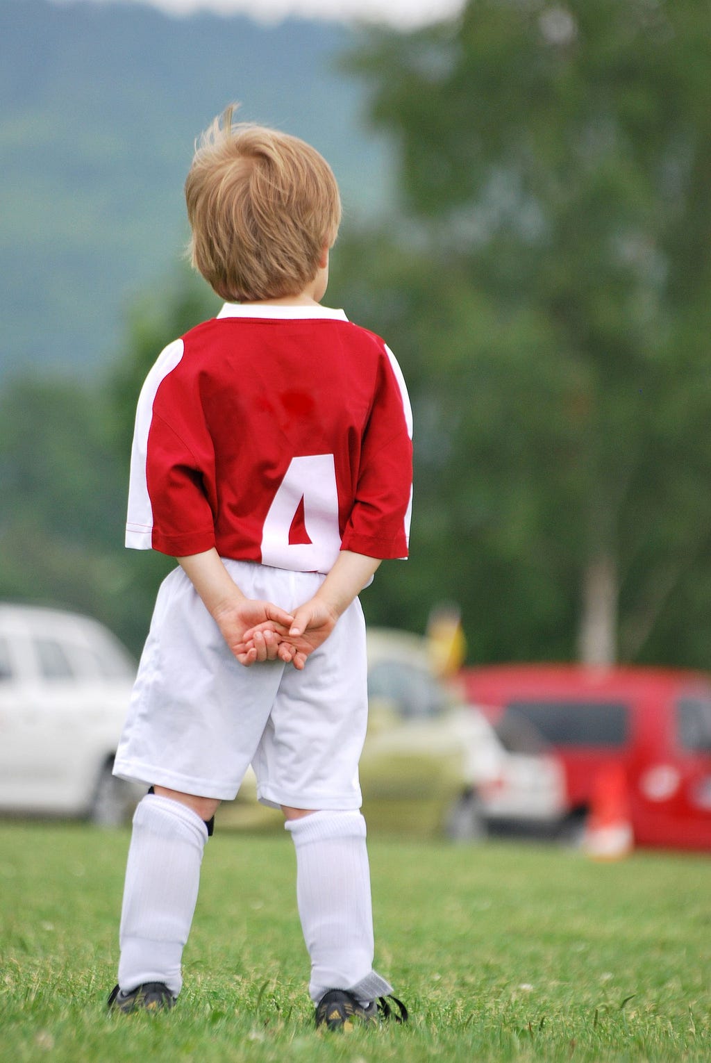 A very young player in a red jersey and white shorts watches the game