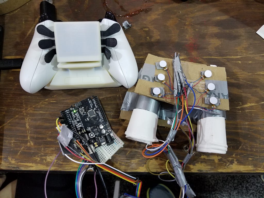 Three prototypes rest on a wooden table. One is made of plastic, another MacGyvered out of cardboard cups and duct tape.