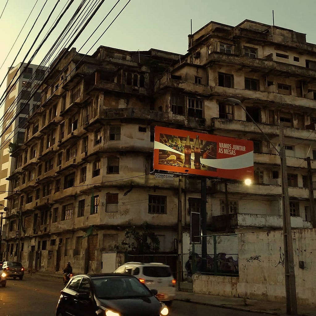 Carros e motos passando pela pista, ao fundo o Edifício São Pedro (abandonado) com um outdoor da Coca-Cola.